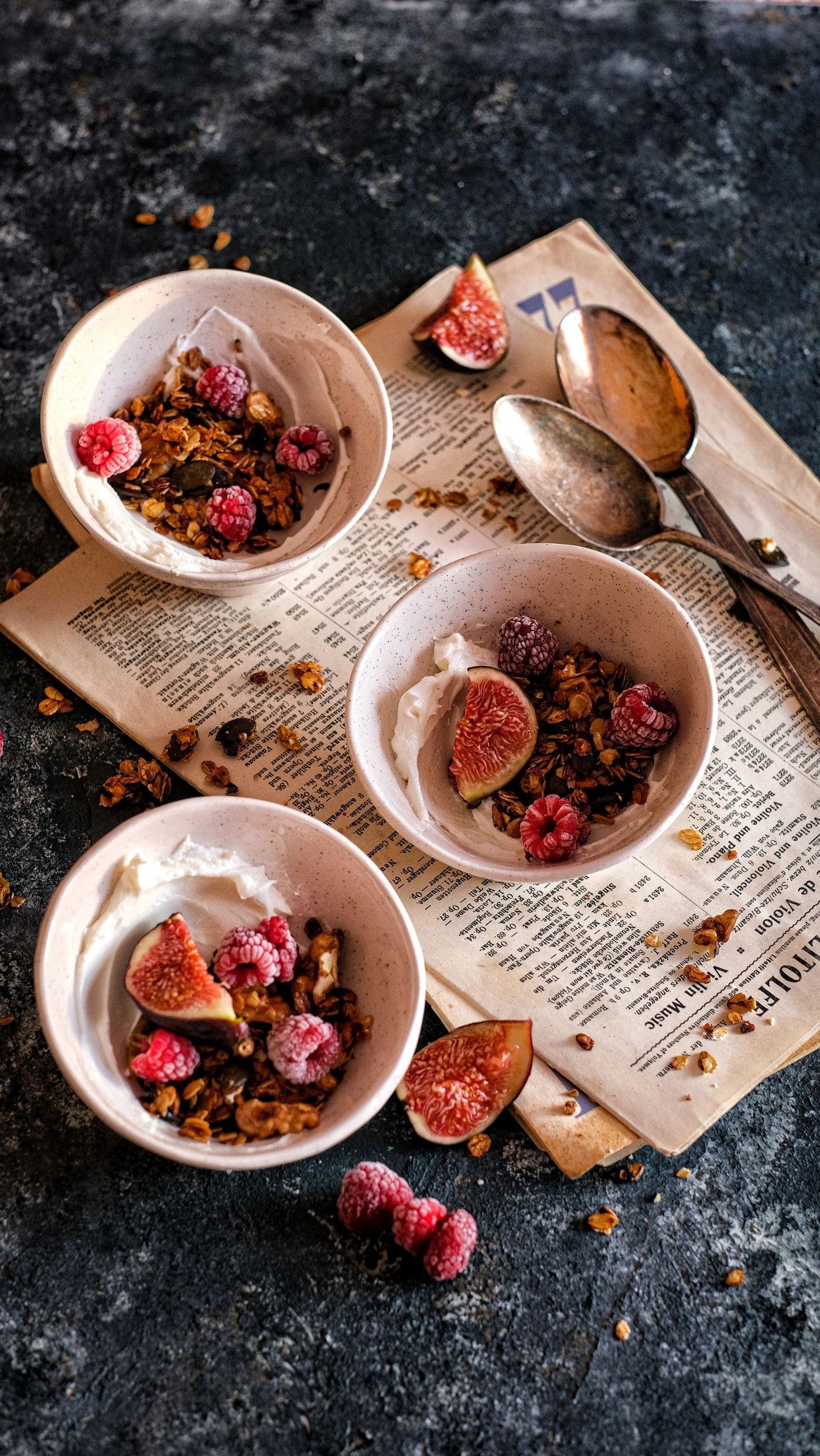 Bowls of granola with yoghurt, fresh figs and raspberries, styled on a dark rustic surface.